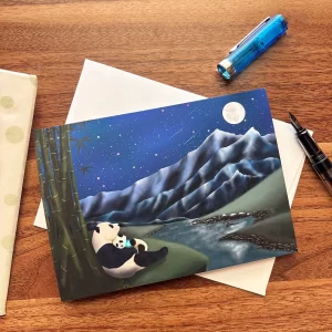 Panda mother and baby night sky greeting card by Tracy Serafini displayed on a wooden desk with envelope and pen